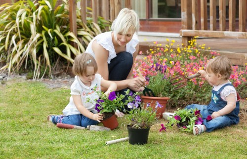 A woman with short blonde hair, wearing a white top, is seated on a well-maintained grassy garden area, engaging with two young children who are planting and watering flowers in small pots. The children, a girl and a boy, are seated on the grass and are dressed in casual clothes, with the girl wearing a white top and blue jeans, and the boy in a blue overall with a white shirt. Around them, there are various flowering plants in pots, displaying vibrant purple, pink, and yellow blooms. In the background, a wooden deck with steps leads up to a house with large windows, and a dense border of ornamental grasses and shrubs forms a natural backdrop. The scene suggests active outdoor gardening and enjoyments of a warm day, with natural sunlight illuminating the garden setup, reflecting typical backyard landscaping and garden maintenance activities in a residential setting.