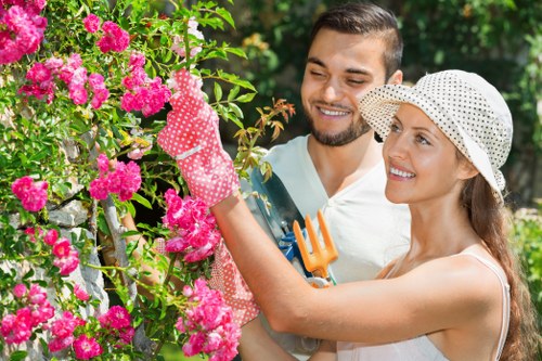 A woman and a man are engaged in gardening activity in a well-maintained outdoor garden. The woman, wearing a white sunhat and dotted gloves, is trimming a vibrant pink flowering shrub with pruning shears, while the man, dressed casually, holds a small hand rake and observes her work. The garden features a lush, green background with a variety of foliage and trees, and the flower bed with bright pink blossoms is situated adjacent to a neatly sheared hedge or shrubbery. The scene depicts a sunny day with natural daylight illuminating the garden, showcasing textures of the dense grass lawn, the soft petals of the flowers, and the greenery of the leaves. This outdoor space is part of a residential garden, likely in Herne Hill or nearby London, reflecting regular garden maintenance activities. Such images support gardening and landscaping services offered by Hedge Trimming Herne Hill, demonstrating professional hedge trimming and shrub care within a typical garden setting.