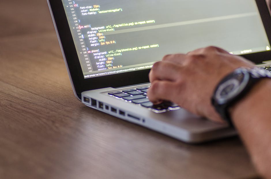 A close-up view of a person's hand using a laptop on a wooden desk, with the screen displaying lines of code in a text editor. The individual is wearing a wristwatch, and the background is blurred, fo